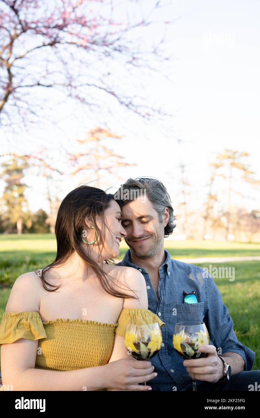 Ein Paar auf einer karierten Picknickdecke im Forest Park mit einer Flasche Wein und roten Knospenbäumen dahinter. Stockfoto