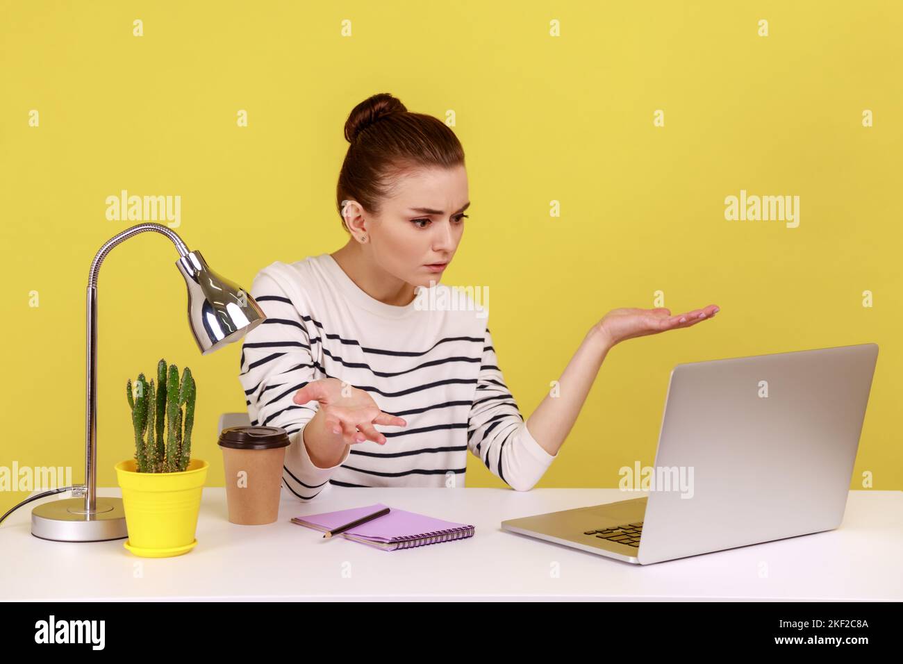 Eine Managerin, die mit einem Laptop am Arbeitsplatz sitzt und mit wütender Empörung auf den Laptop-Bildschirm blickt und Konflikte mit Kollegen hat. Studio-Innenaufnahme isoliert auf gelbem Hintergrund. Stockfoto