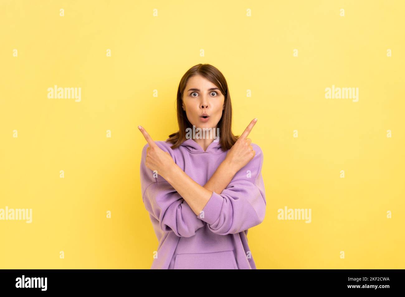 Porträt einer überraschten dunkelhaarigen Frau, die mit gekreuzten Händen steht und auf beiden Seiten den Raum kopieren soll, mit einem violetten Hoodie. Innenaufnahme des Studios isoliert auf gelbem Hintergrund. Stockfoto