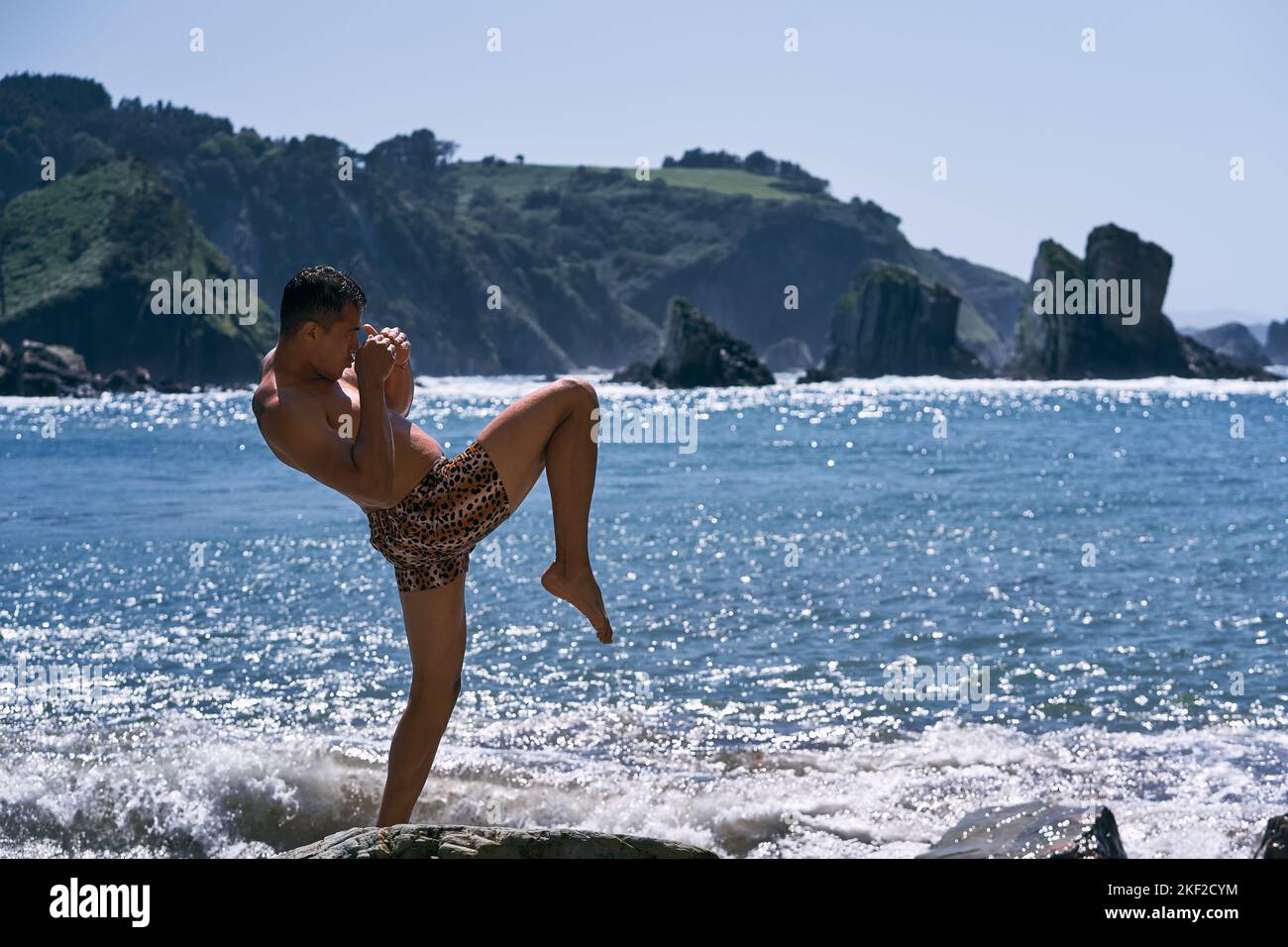 lateinischer junger Mann, der einen Tiger im Profil trägt, seine Arme gefaltet und dabei nasse Kampfkunsthaltungen mit einem Bein im Meerwasser in der Nähe des macht Stockfoto