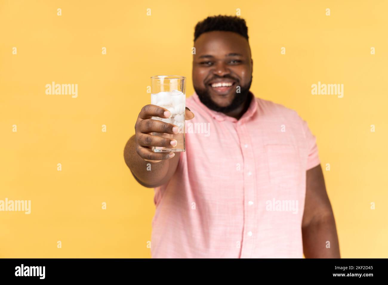 Porträt eines positiven glücklichen optimistischen Mannes in einem rosa Hemd, der ein Glas Wasser mit Eis aushält und Ihnen kaltes Getränk anbietet. Innenaufnahme des Studios isoliert auf gelbem Hintergrund. Stockfoto