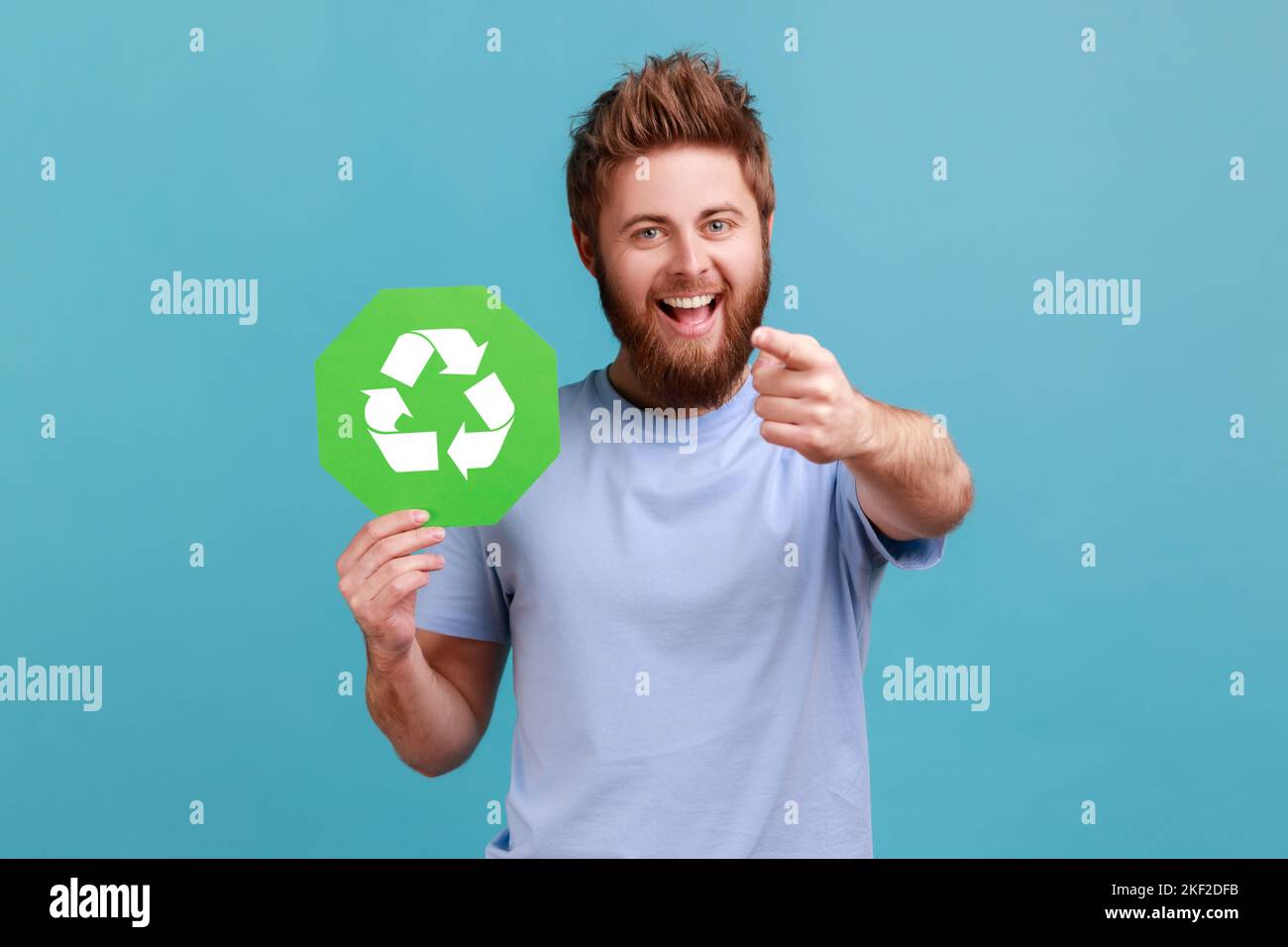 Porträt eines äußerst glücklichen, aufgeregt bärtigen Mannes mit grünem Recycling-Schild, Umweltschutz, Ökologie-Konzept, zeigt auf die Kamera. Innenaufnahme des Studios isoliert auf blauem Hintergrund. Stockfoto