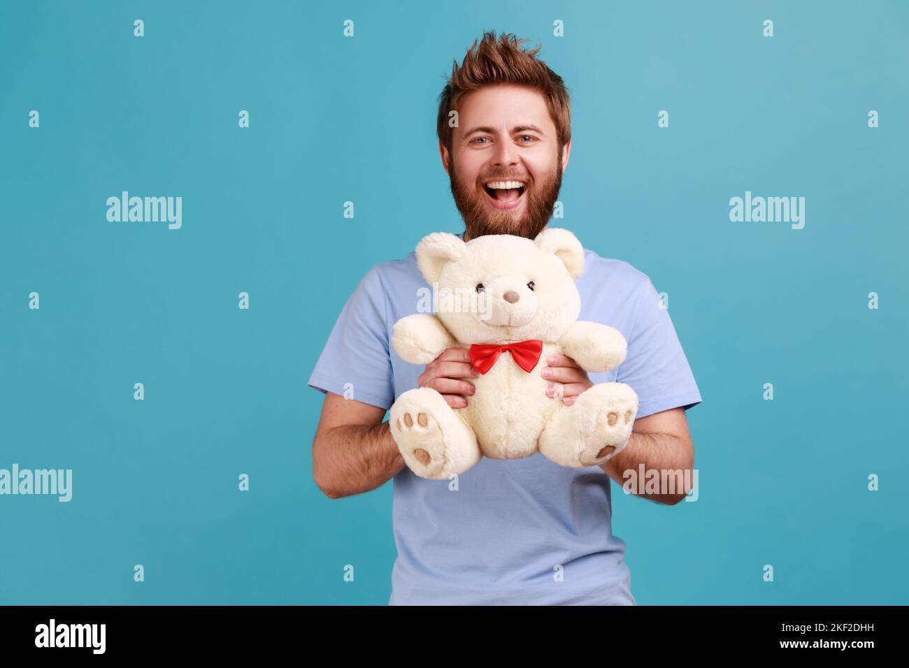 Porträt eines glücklichen optimistischen, gutaussehenden bärtigen Mannes, der mit einem weißen, weichen Teddybären auf die Kamera blickt und positive Emotionen ausdrückt. Innenaufnahme des Studios isoliert auf blauem Hintergrund. Stockfoto