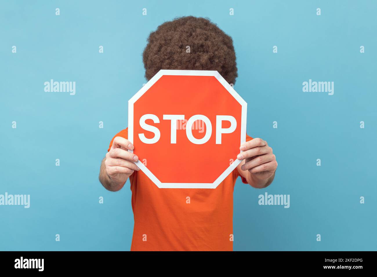 Porträt eines Mannes mit Afro-Frisur, der ein orangefarbenes T-Shirt-bedecktes Gesicht mit Stopp-Symbol trägt, anonyme Person mit rotem Verkehrsschild, Warnung vor dem Start. Innenaufnahme des Studios isoliert auf blauem Hintergrund. Stockfoto