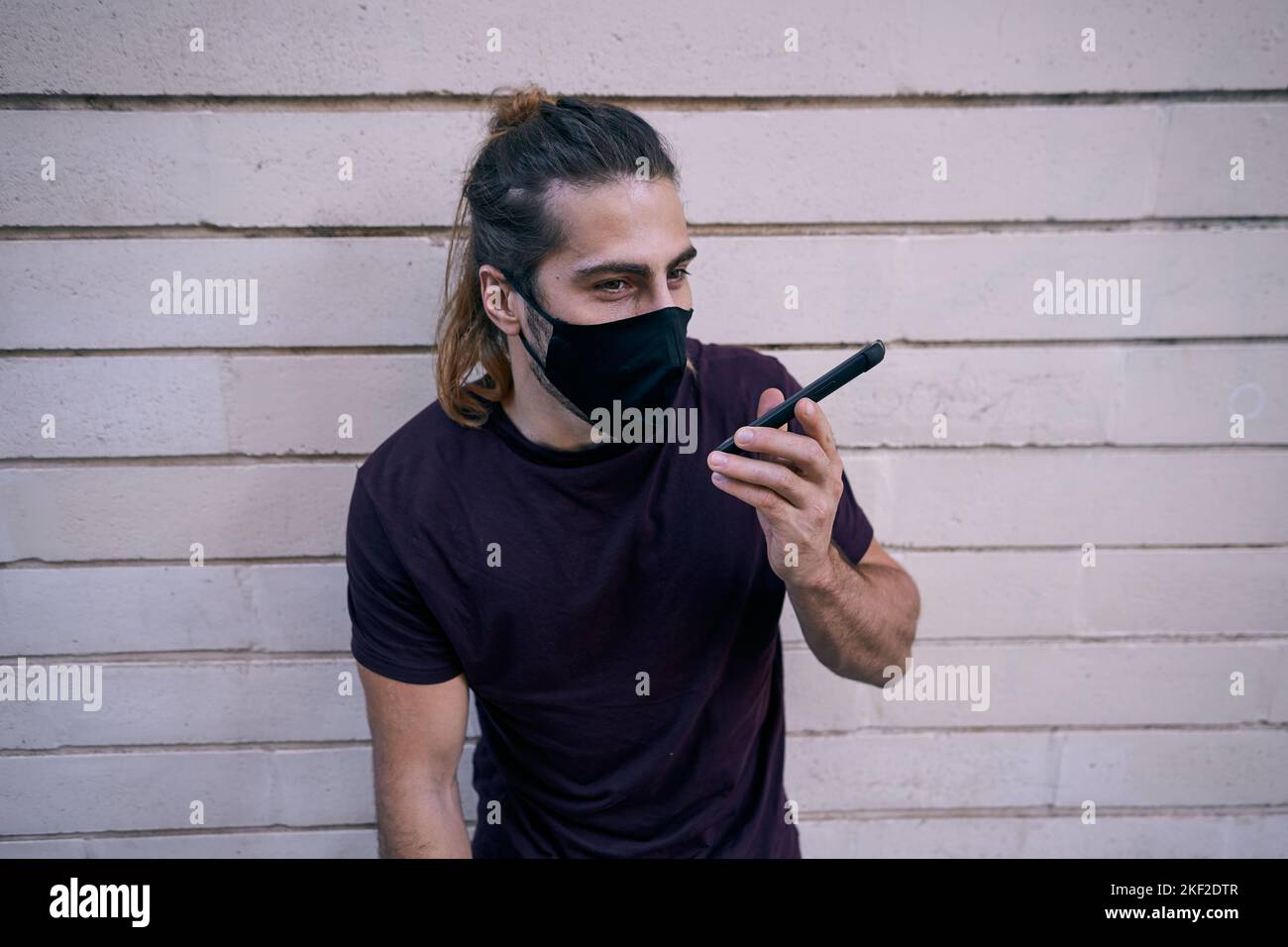 kaukasischer Kerl mit langen Barthaar, das in einem schwarzen T-Shirt an der Wand gelehnt steht und auf dem Smartphone mit Gesichtsmaske spricht Stockfoto