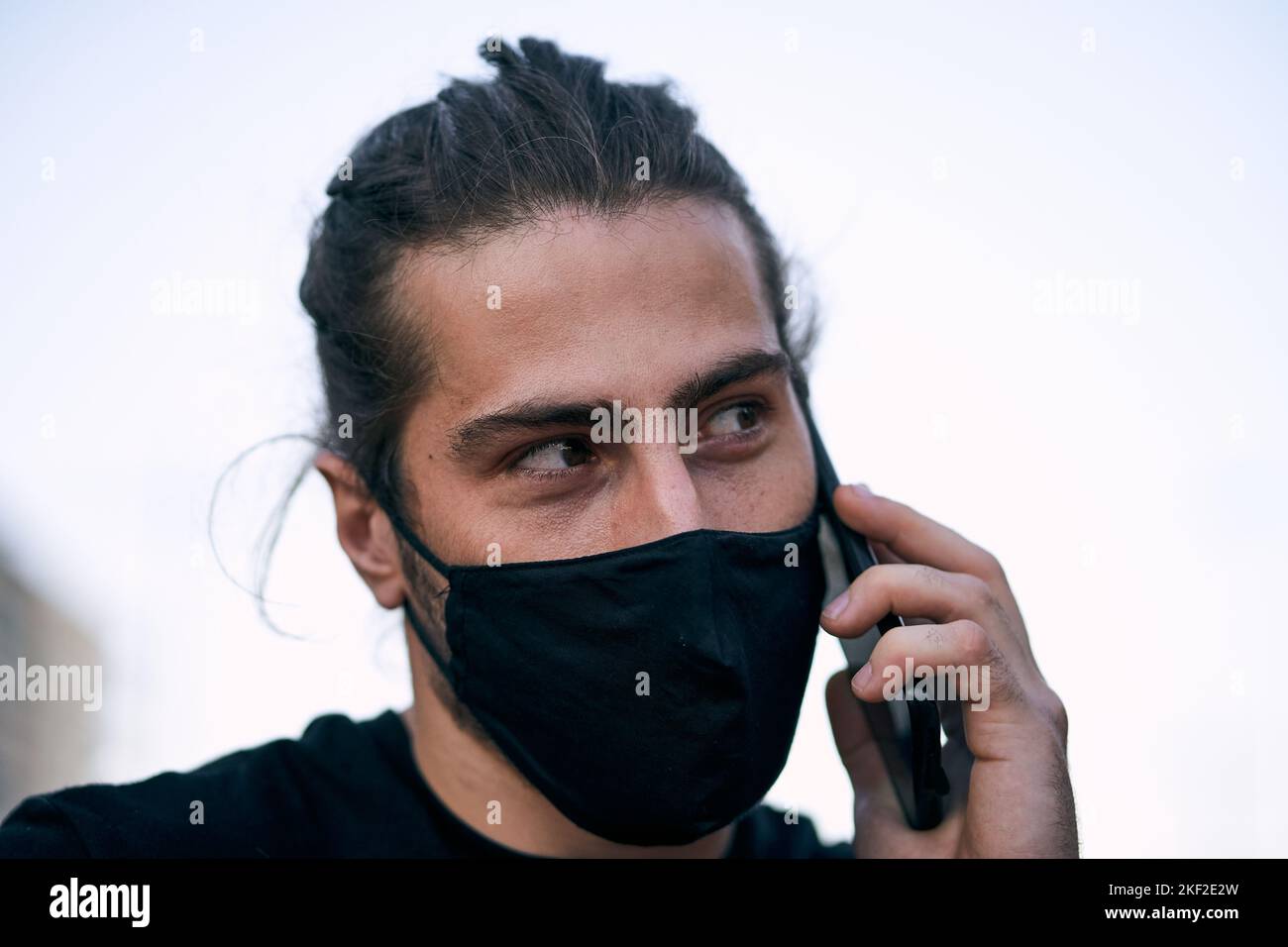 der kaukasische Typ mit schwarzem T-Shirt zerzauste lange Haare mit Gesichtsmaske auf der Straße und sprach auf seinem Smartphone in der Stadt Stockfoto