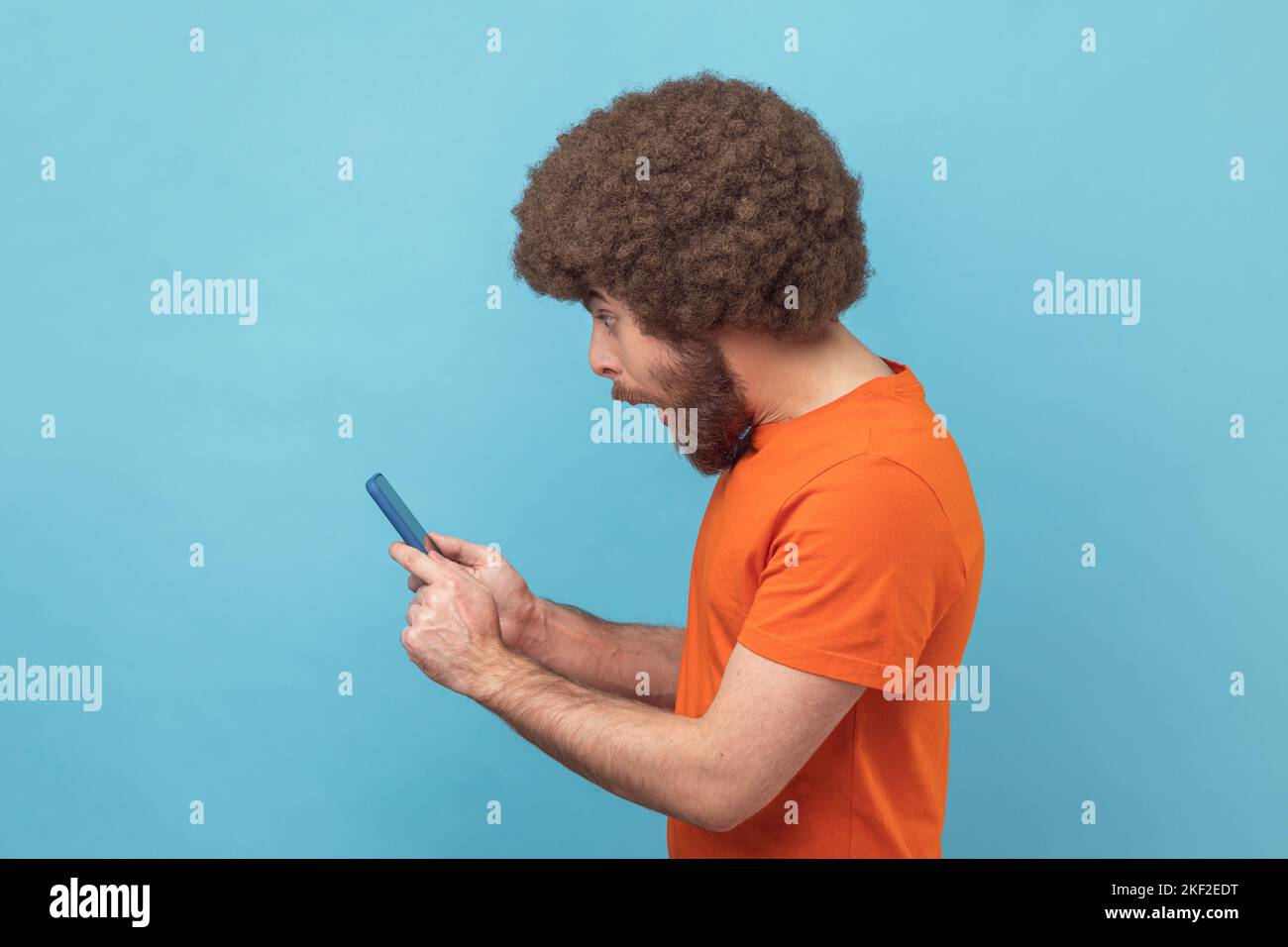 Seitenansicht eines staunenden Mannes mit Afro-Frisur, der ein orangefarbenes T-Shirt trägt und einen Beitrag über ein Mobiltelefon im sozialen Netzwerk liest und überrascht chattet. Innenaufnahme des Studios isoliert auf blauem Hintergrund. Stockfoto