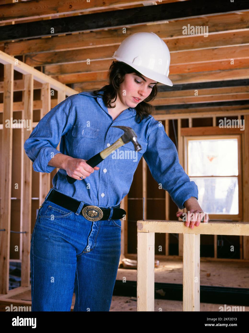 1970S DIY BRÜNETTE FRAU ARBEITET ALS ZIMMERMANN AUF DER BAUSTELLE MIT HAMMER UND NÄGEL TRAGEN DENIM ARBEITSKLEIDUNG HARD HAT - KS15247 VRE001 HARS BAUMWOLLE LIFESTYLE ZUFRIEDENHEIT NAGEL FRAUEN VERTIKALE ARBEITSPLÄTZE WEBSITE ZIMMERMANN KOPIEREN RAUM HALBE LÄNGE PERSONEN INSPIRATION NÄGEL VERTRAUEN DENIM WOMAN'S BRÜNETT BLAU KRAGEN BERUF SCHÜTZEN STRUKTUR ARBEITER STRAHL MANUELLEN SCHUTZ HARD HAT UND WOMAN'S BEFREIUNG FRAUEN'S RECHTE LIB LOW-ANGLE WOMAN'S RIGHTS WORKFORCE WOMEN'S LIB WOMEN'S LIBERATION LABOUR PRIDE RIGHTS WORKERS OPPORTUNITY HARDHAT-BERUFE MIT HÄMMERN, DIE KONSTRUKTION NAGELN STANDORT Stockfoto