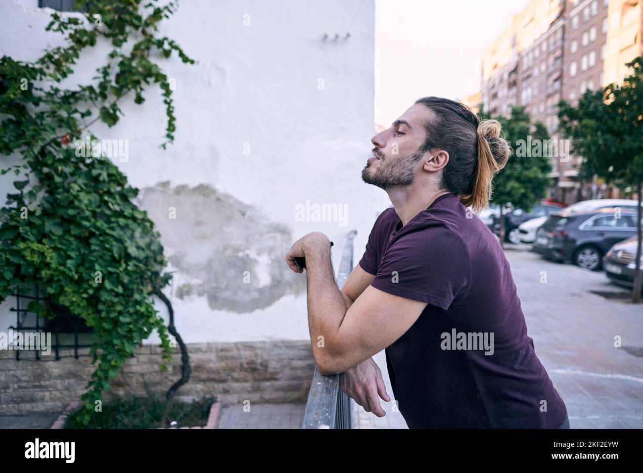 kaukasischer Kerl, der in einem schwarzen T-Shirt steht und dampfenden Rauch ausatmet, der sich auf das Metallgeländer einer Stadtstraße stützt Stockfoto