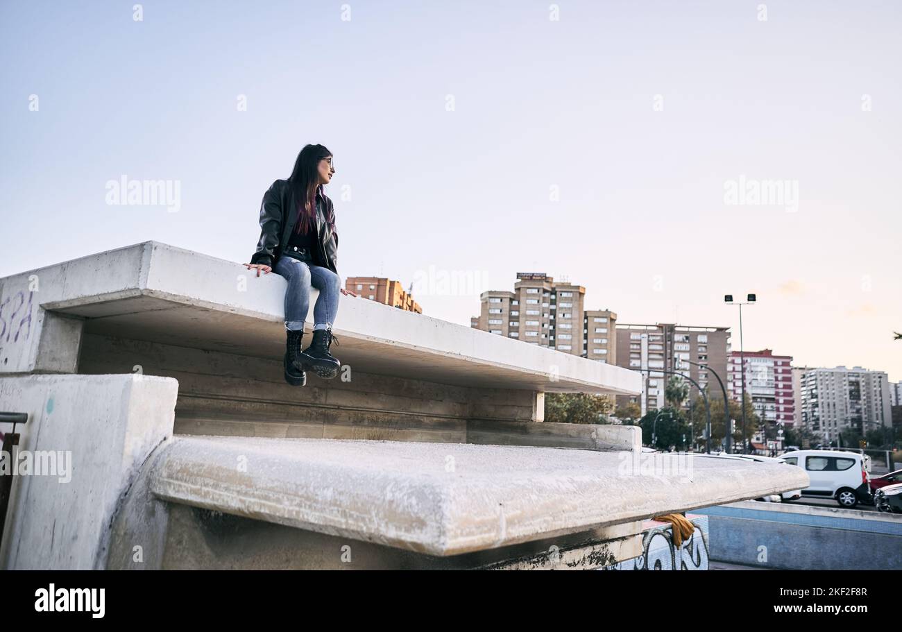 latina Mädchen in zerrissenen blauen Hosen Pelz Jacke Brille Blick auf die Stadt ruhig und entspannt auf der Betonwand sitzen Stockfoto