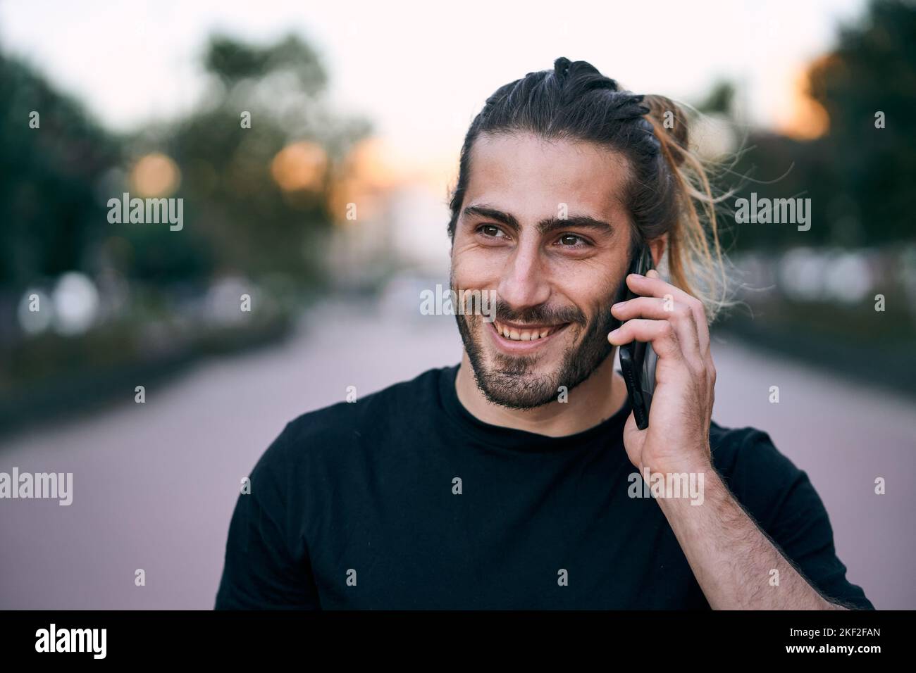 das schwarze T-Shirt des kaukasischen Mannes zerzauste lange Haare mit einem lächelnden Bart, der auf der Straße auf seinem Smartphone auf dem Stadtboulevard stand und sprach Stockfoto