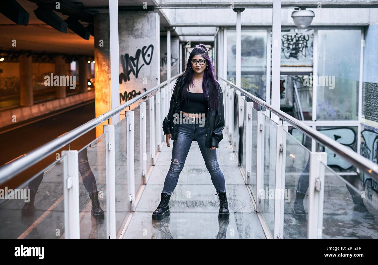 latina Mädchen mit langen Haaren Brünette Brille Pelzjacke zerrissene blaue Jeans und große schwarze Stiefel stehend Blick auf die Kamera mit Beinen auf Glas gespreizt Stockfoto