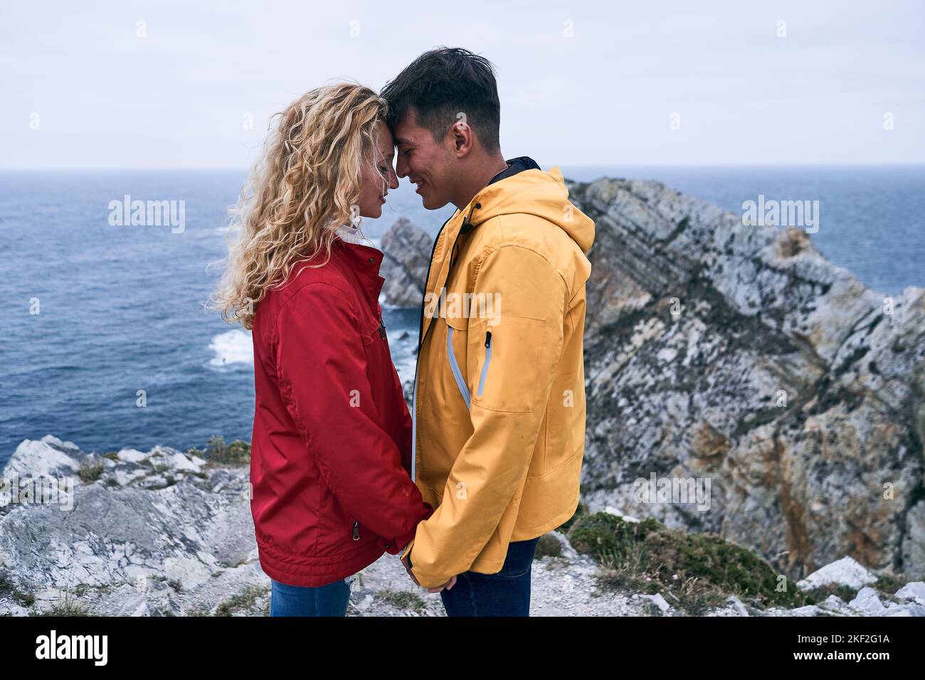 lateinischer Junge und blondes kaukasisches Mädchen im Regenmantel stehen im Profil gegenüber einander mit Stirn in Kontakt lächelnd die Hände vor dem halten Stockfoto