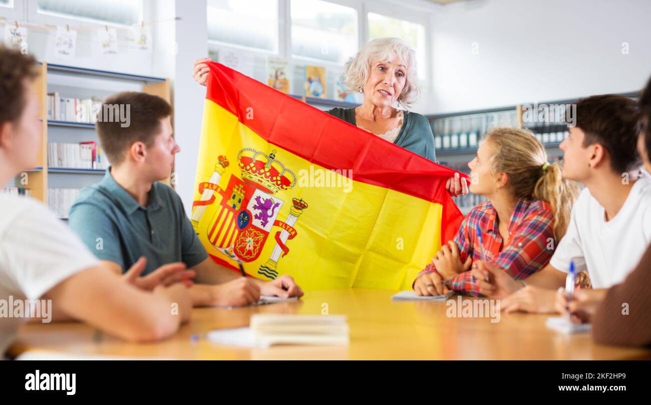 Reife Lehrerin zeigt Teenagern die Flagge Spaniens und spricht während des Unterrichts an der Bibliotheksschule über dieses Land Stockfoto