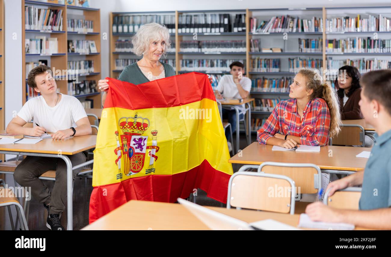 Reife Lehrerin zeigt Teenagern die Flagge Spaniens und spricht während des Unterrichts an der Bibliotheksschule über dieses Land Stockfoto