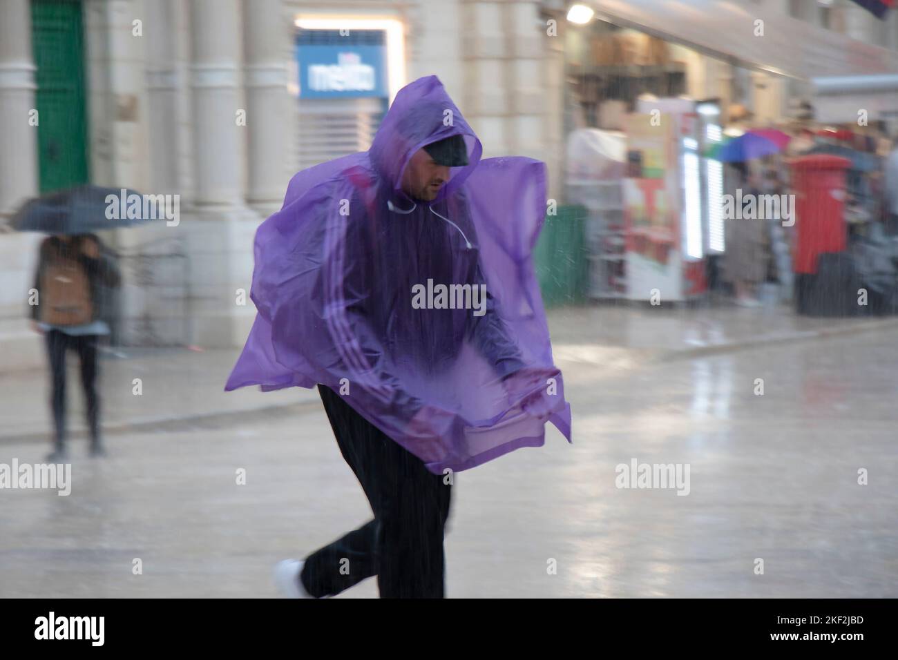 Valletta, Malta - 12. November 2022: Ein Mann mit purpurfarbenem Notregen-Poncho läuft, um sich vor dem fallenden Regen zu verstecken, in Bewegung verschwommen Stockfoto