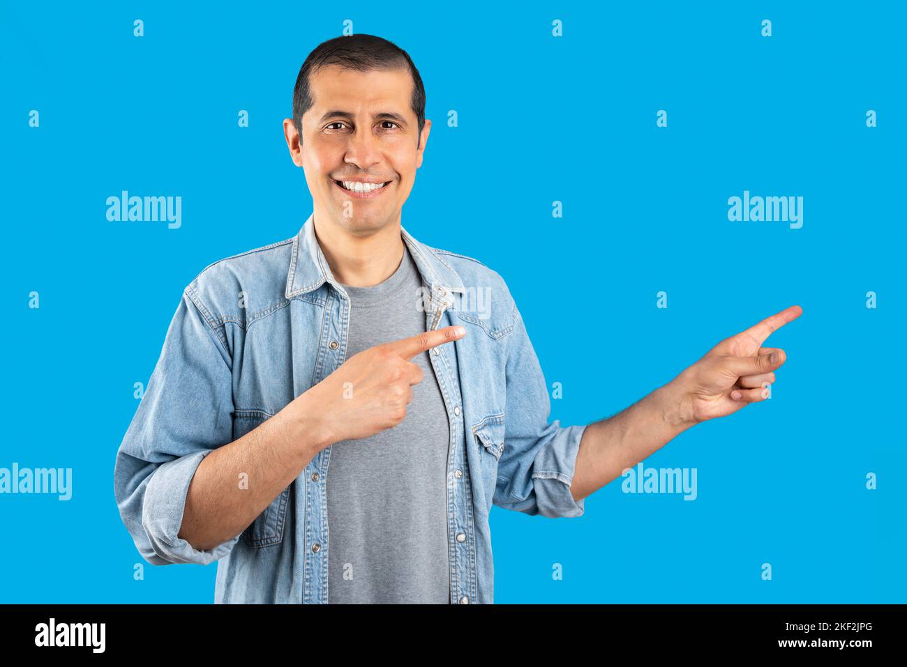 Studioaufnahme eines hispanischen Mannes in Jeanshemd, der lächelnd auf einem isolierten blauen Hintergrund stand und mit zwei Händen und auf die Kamera zeigte Stockfoto