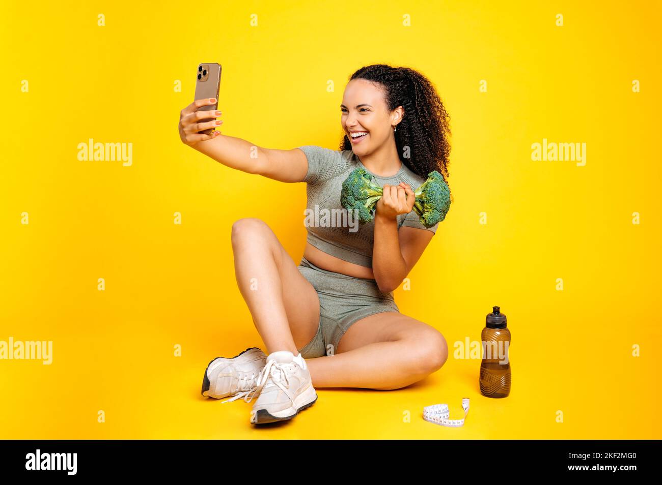 Fröhliche lustige gemischte Rasse lockige Frau im Sport-Outfit, nimmt Selfie auf dem Smartphone mit Brokkoli Hantel, während sie auf isolierten orangefarbenen Hintergrund sitzen, lächeln, Spaß haben, emotionalen Gesichtsausdruck Stockfoto