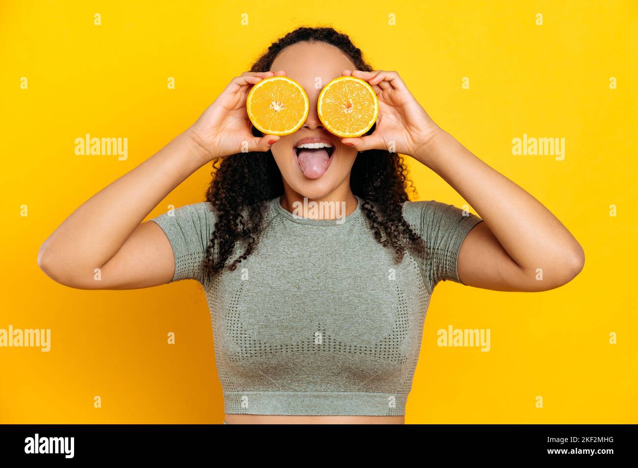Fröhliche liebliche brasilianische oder latino junge lockige haarige Frau, in Sport-Outfits, stehend auf isoliertem orangefarbenem Hintergrund, hält zwei Hälften orange in den Händen neben den Augen, zeigt Zunge, lächelnd Stockfoto