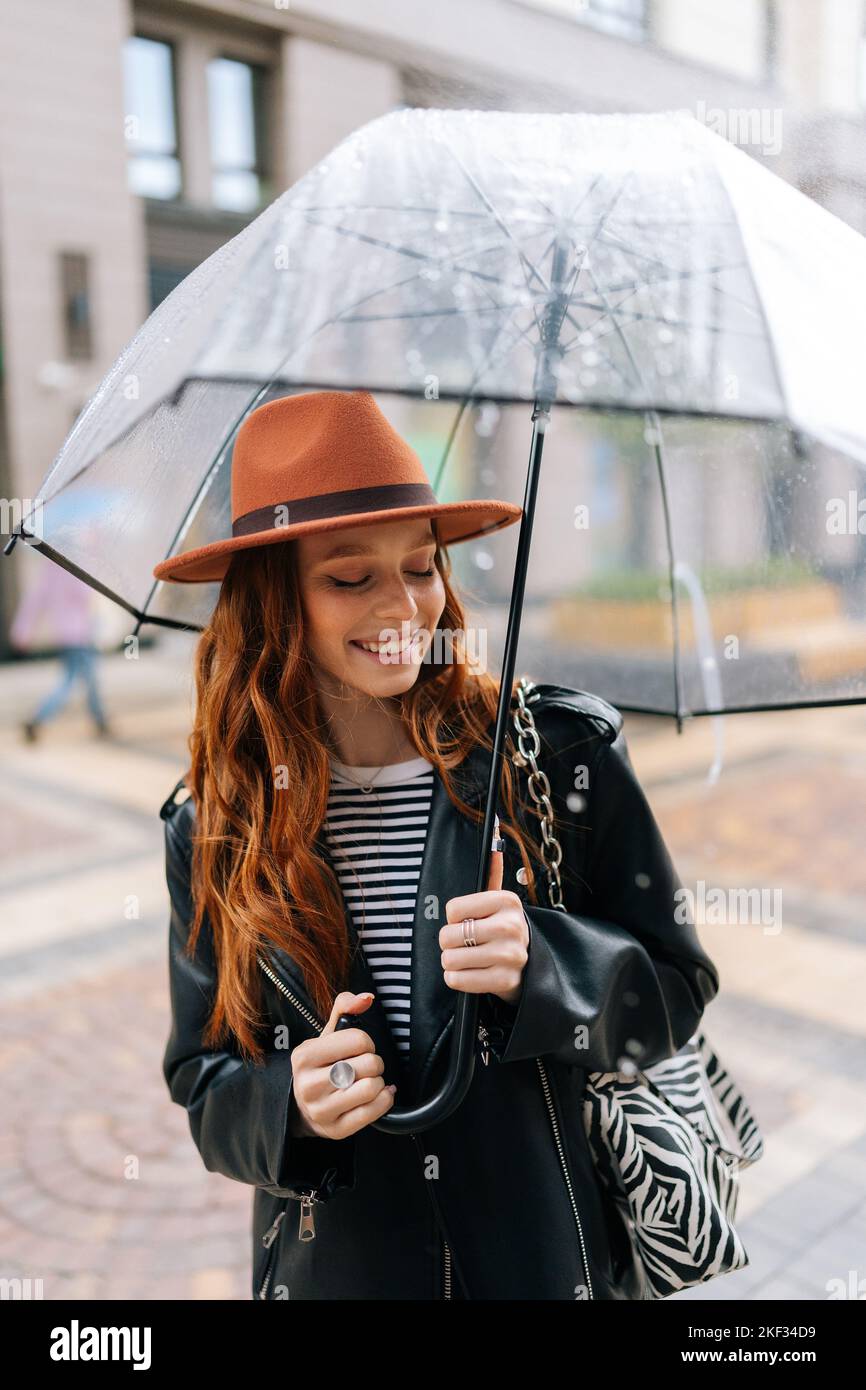 Vertikale Aufnahme einer fröhlichen jungen Frau im Hut, die mit  transparentem Regenschirm auf der Straße der Stadt steht und das  regnerische Wetter im Freien genießt und lächelnd nach unten blickt  Stockfotografie -
