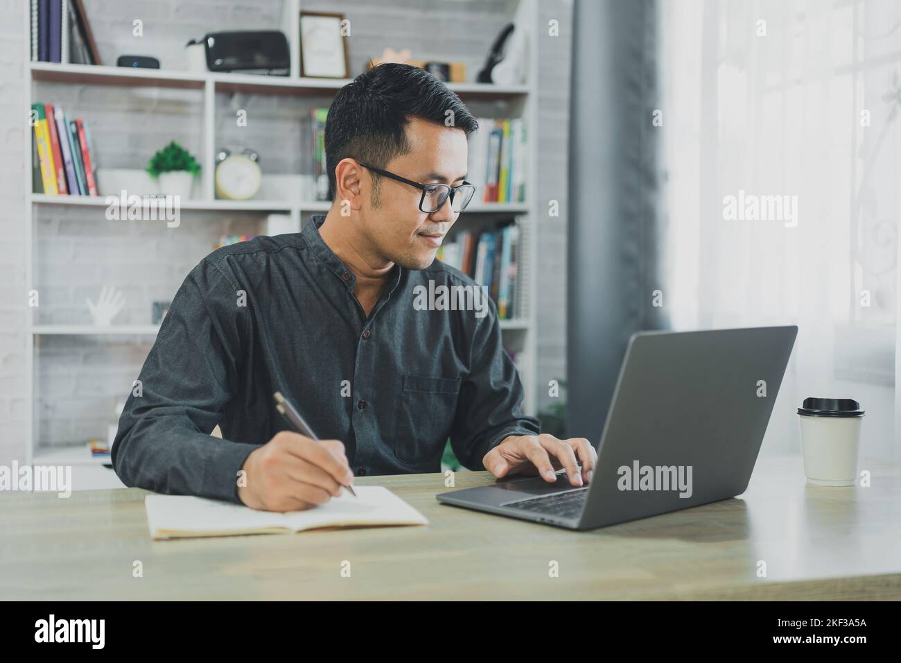 Der freiberufliche Mann aus Asien trägt eine Brille und lächelt beim Tippen auf dem Laptop und schreibt zu Hause Notizen im Notebook. Unternehmer Mann, der für Unternehmen bei l Stockfoto