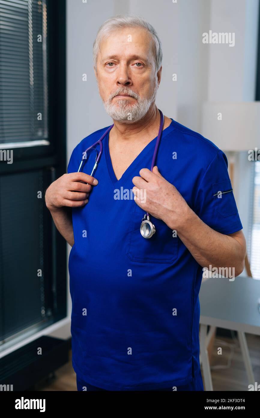Vertikales Porträt eines traurigen überarbeiteten älteren männlichen Arztes in dunkelblauer medizinischer Uniform, der im Krankenhausbüro steht und die Kamera anschaut. Stockfoto