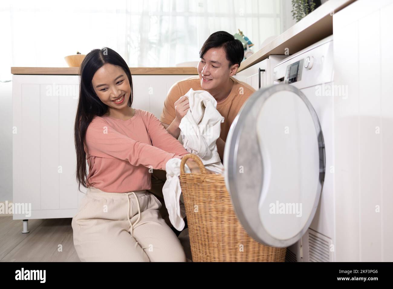 Junge Asiaten heirateten glückliches Paar lächelnd und machen Wäsche zu Hause. Freund und Freundin, die ihre Kleidung vor dem Laden der Waschmaschine zusammenlegen Stockfoto