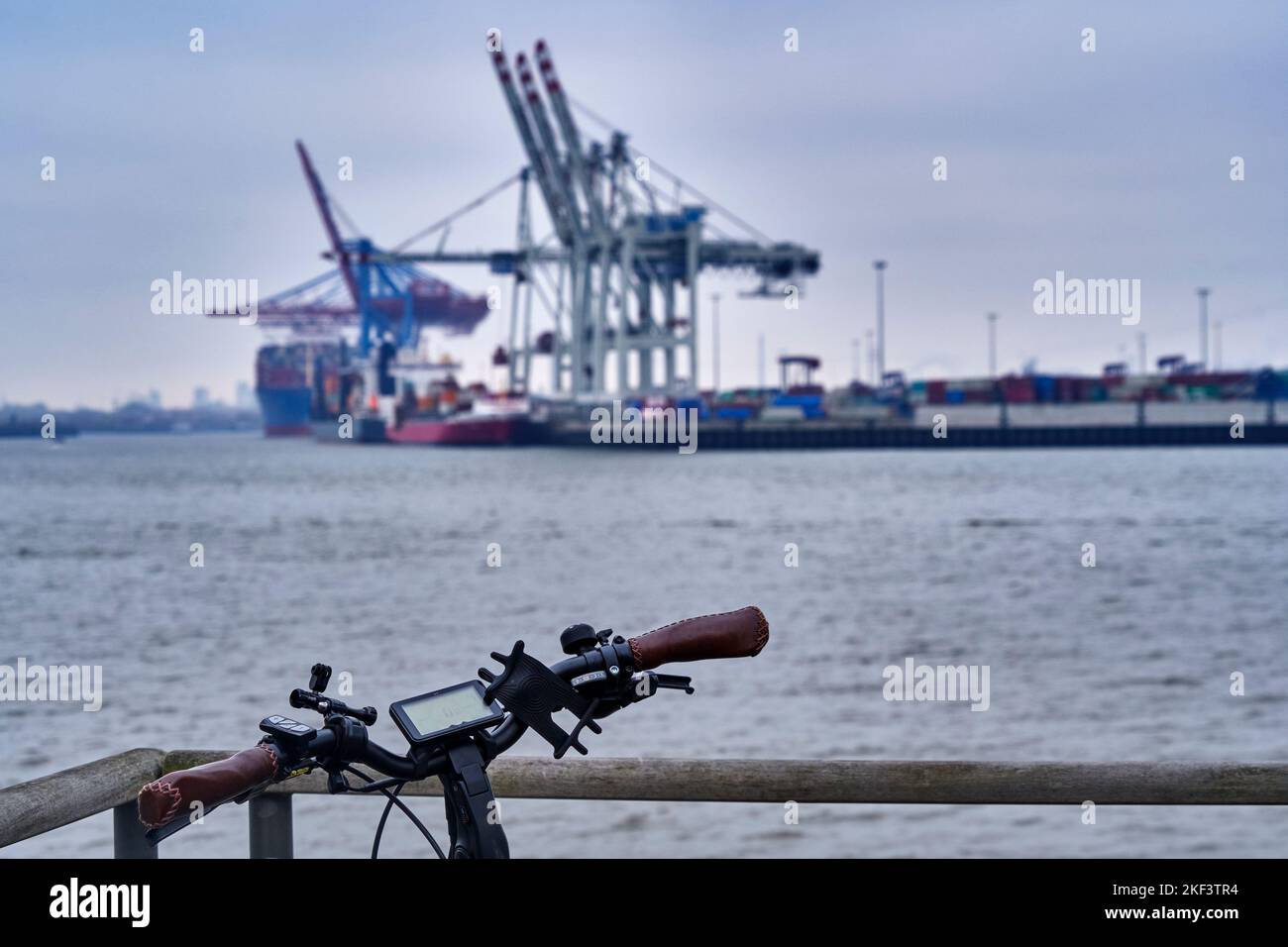 Lenker eines schwarzen Elektrofahrrads, das in Hamburg gegen eine Brüstung gelehnt ist, verschwommener Hintergrund mit der Elbe und Containerkranen, selektiver FOC Stockfoto