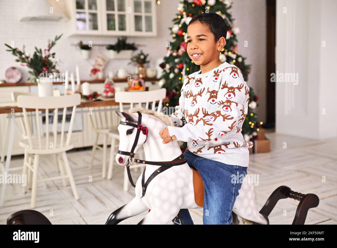 Kleiner afroamerikanischer Junge in einem grauen Weihnachtspullover mit Hirschen lächelt, während er auf einem Schaukelpferd vor dem Hintergrund eines Weihnachtsfestes sitzt Stockfoto