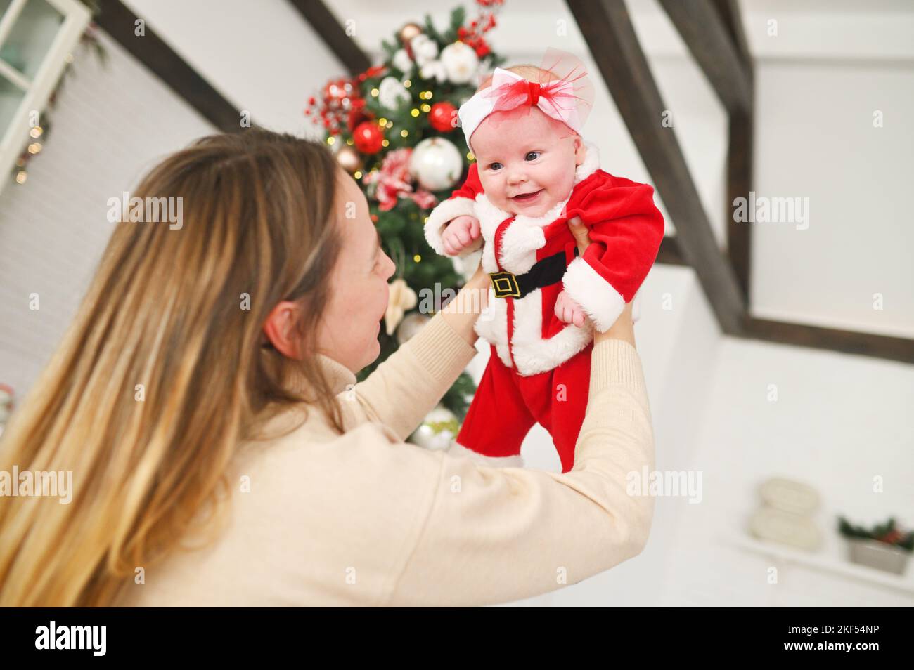 Mama hält ein Baby in einem weihnachtsmann-Kostüm auf dem Hintergrund eines weihnachtsbaums in den Armen. Stockfoto