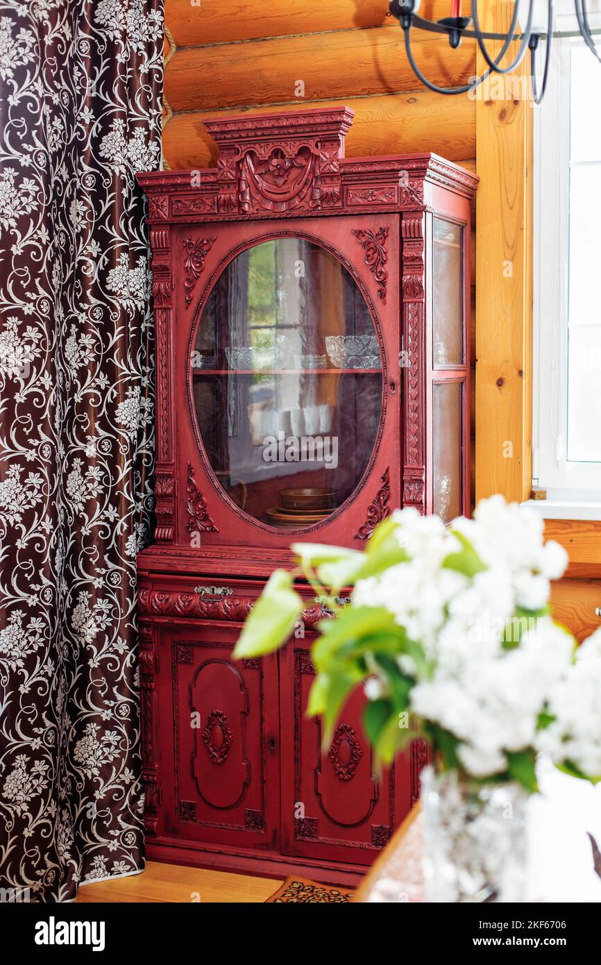 Alter antiker roter Holzschrank mit geschnitzten Dekorationen in der Ecke der Blockhütte in der Nähe eines braunen Vorhangs mit Blumenmustern. Stockfoto