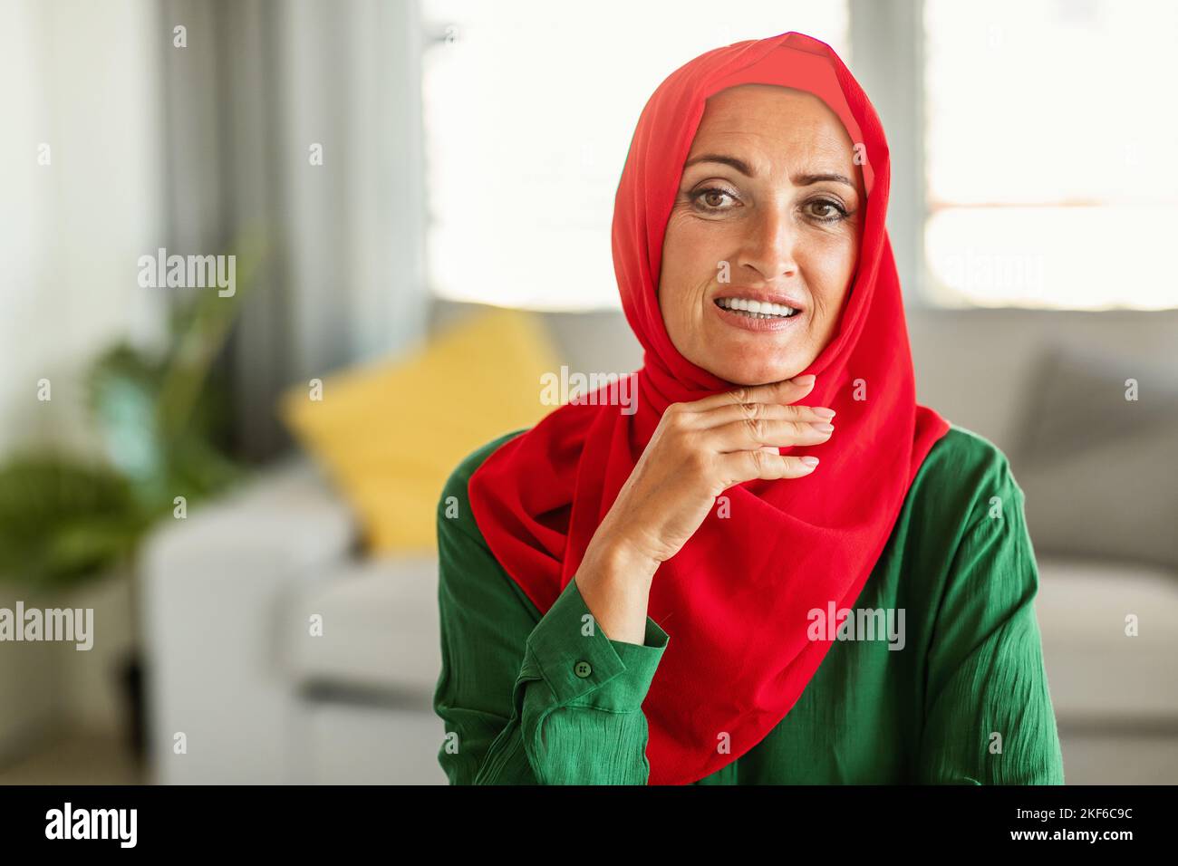 Glückliche muslimische Frau mittleren Alters, die roten Hijab trägt, vor der Kamera posiert und lächelt und zu Hause im Wohnzimmer sitzt Stockfoto