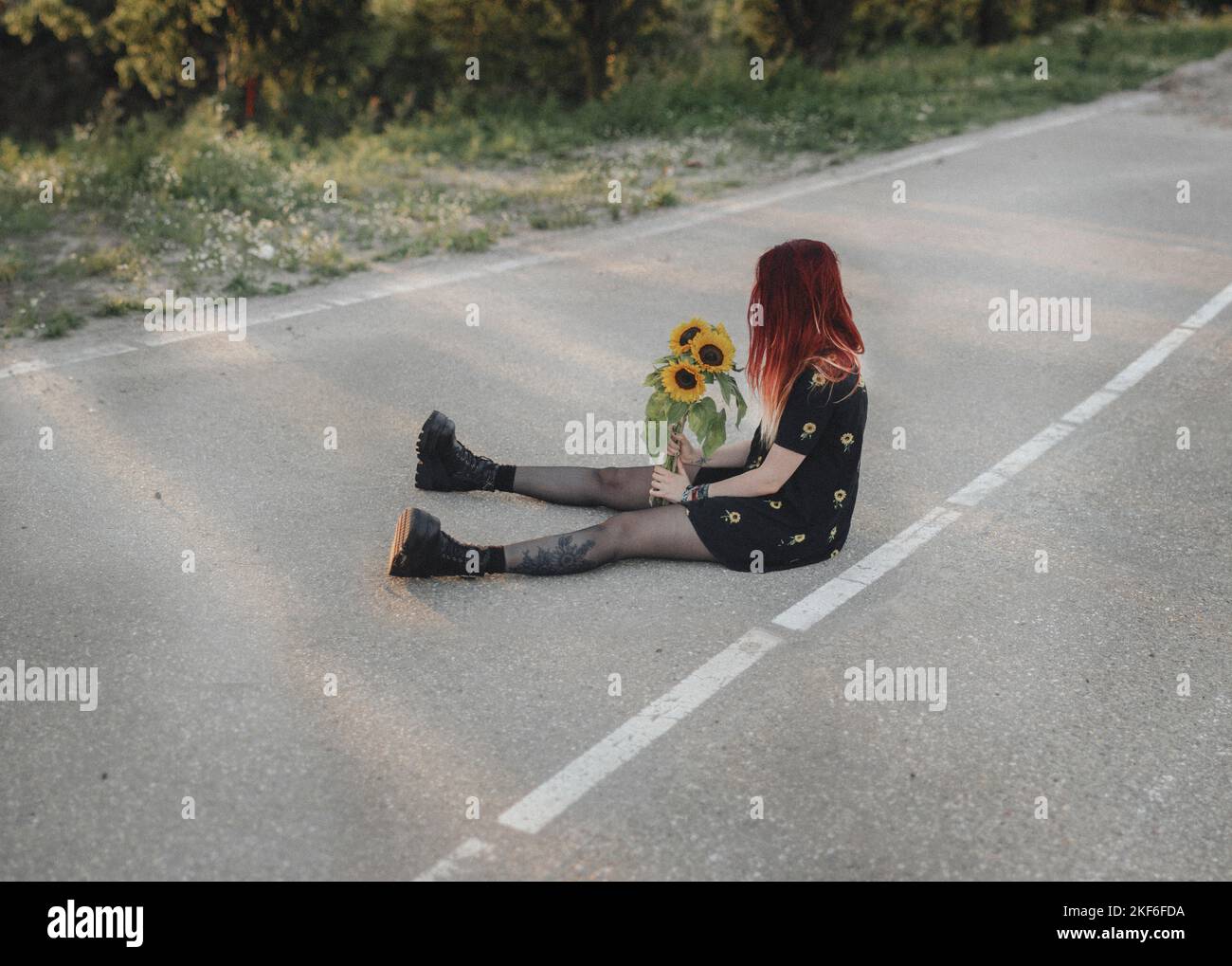 Junge Frau mit roten Haaren, die Sonnenblumen hält und auf einer leeren Straße sitzt Stockfoto