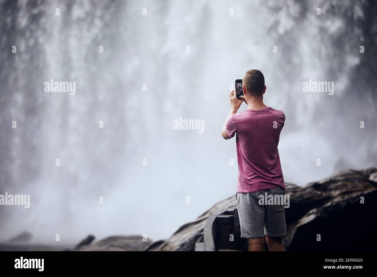 Rückansicht des Touristen beim Fotografieren mit dem Handy. Mann vor hohem Wasserfall in Kambodscha. Stockfoto