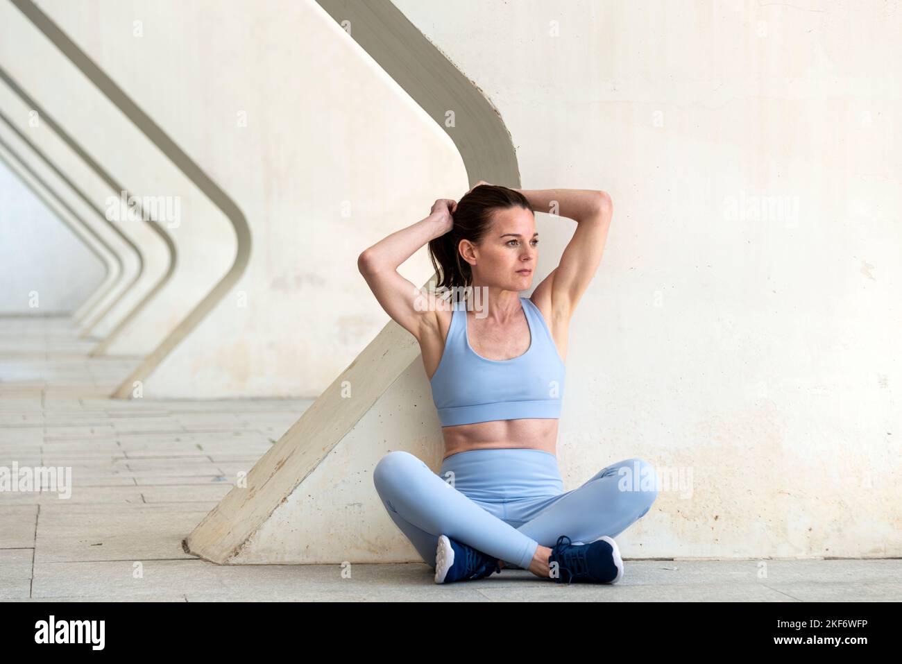 Eine sportliche Frau schnürt sich vor dem Laufen die Haare und trainiert draußen in einer urbanen Umgebung. Sitzend mit gekreuzten Beinen. Stockfoto