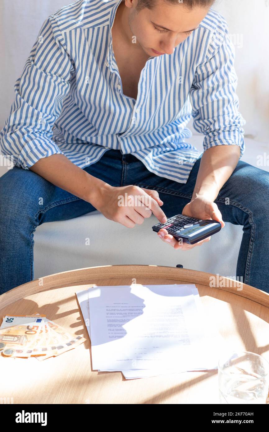 Vertikale Ansicht eines jungen Mannes, der auf der Couch sitzt und mit beutlichem Sonnenlicht Berechnungen und Papierkram für den Haushalt macht Stockfoto
