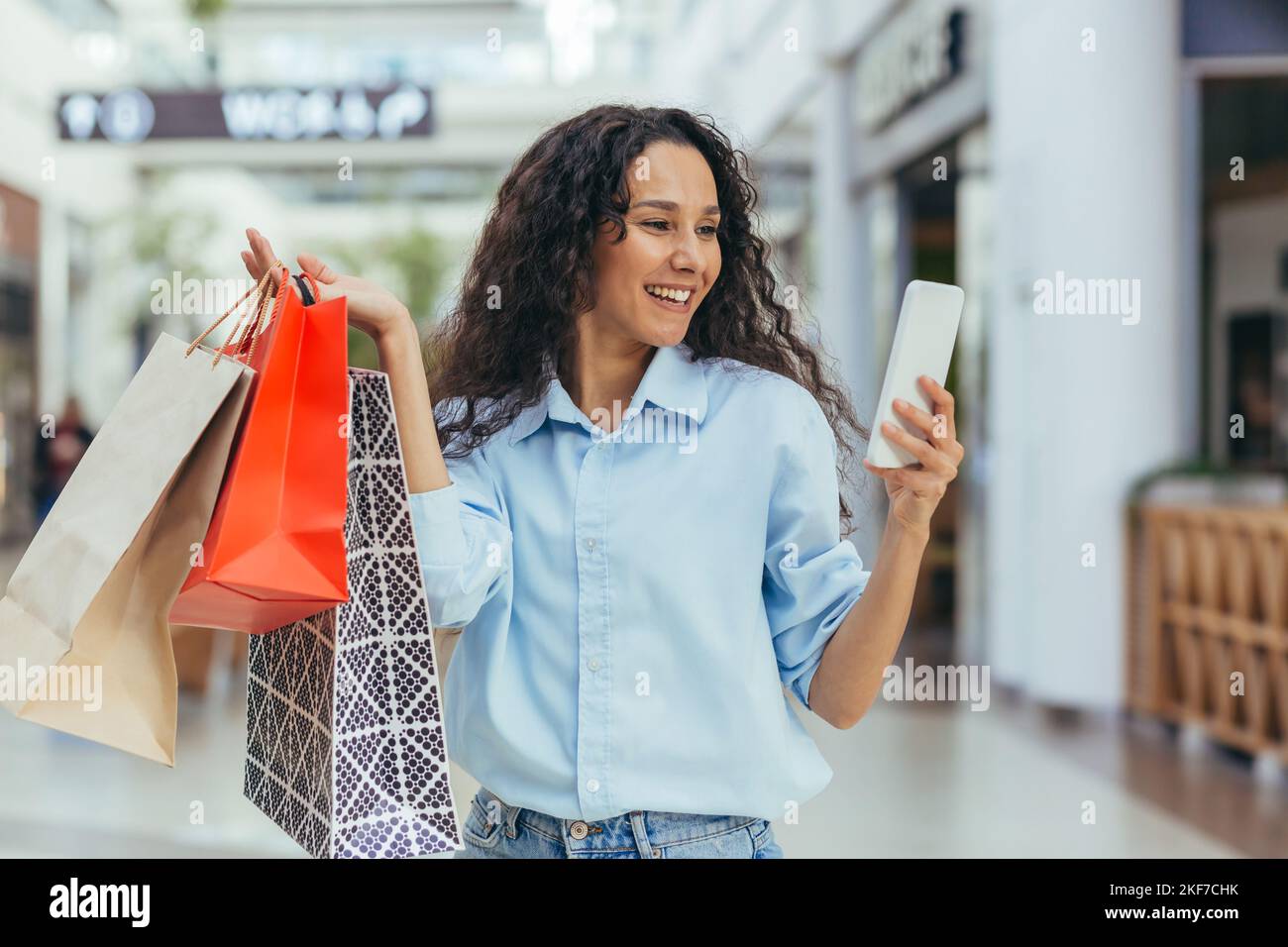 Eine attraktive junge lateinamerikanische Frau kauft in einem Einkaufszentrum ein und hält bunte Taschen in ihren Händen. Nutzt das Telefon, macht ein Selfie, lächelt. Stockfoto