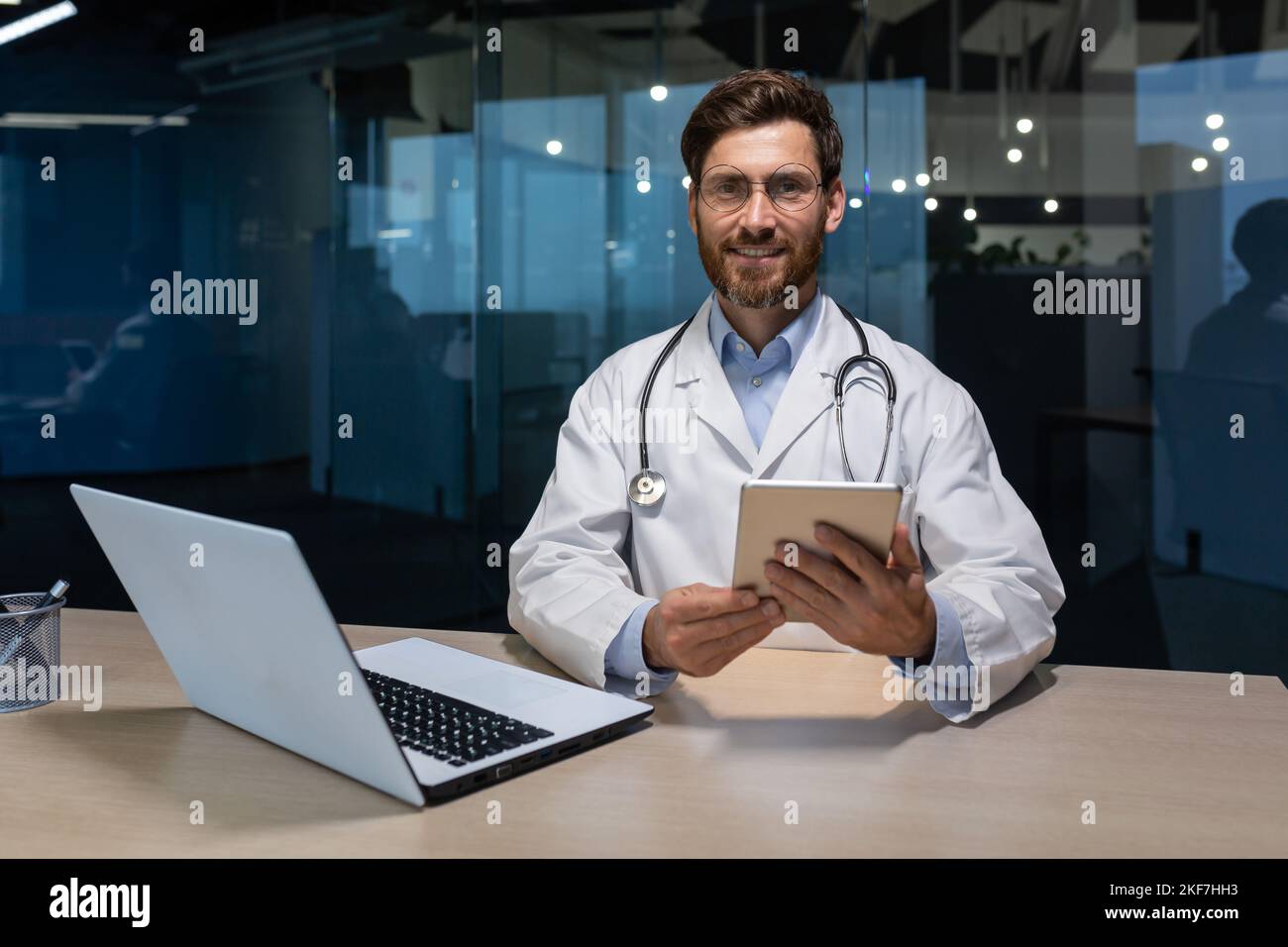 Porträt eines jungen hübschen männlichen Arztes, der mit einem Laptop in einem Büro in einem Krankenhaus an einem Tisch sitzt und arbeitet. Hält ein Tablet in den Händen, blickt auf die Kamera und lächelt. Stockfoto