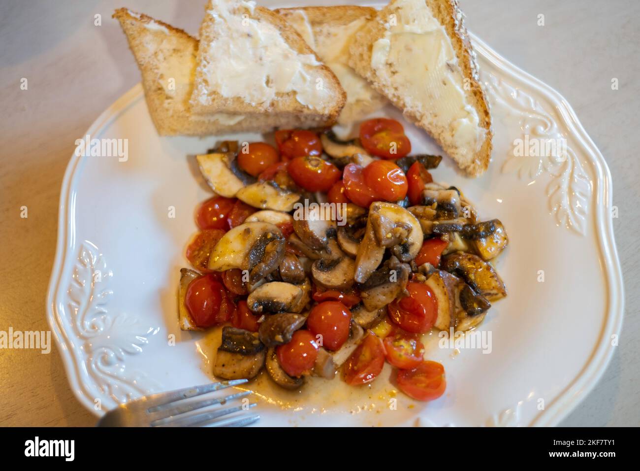Gesundes Gemüseessen mit Pilzen, Kirschtomaten und Knoblauch, gebraten oder sautiert und auf einem weißen Teller mit Brot und Butter serviert. Stockfoto