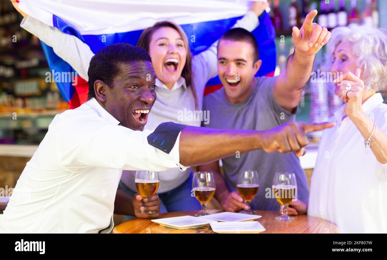 Fröhliche Freunde unterschiedlichen Alters trinken Bier und feiern den Sieg des russischen Teams in der Nachtbar Stockfoto