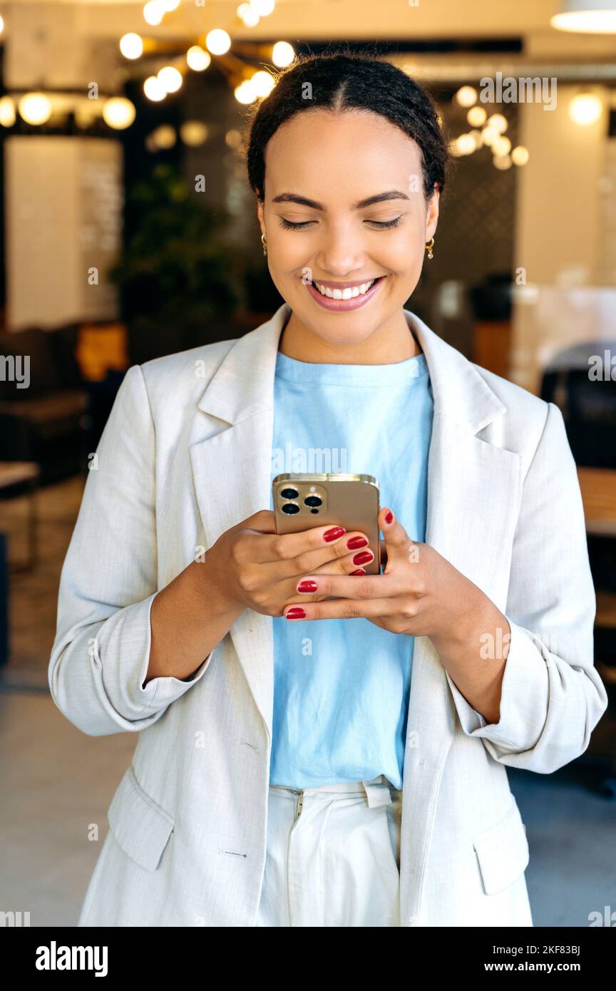 Online-Messaging. Fröhliche Geschäftsfrau mit gemischter Rasse, Managerin eines Unternehmens, die in ihrem modernen kreativen Büro steht, nutzt ihr Smartphone, Messaging in sozialen Medien, beantwortet E-Mails, tippt Text, lächelt Stockfoto