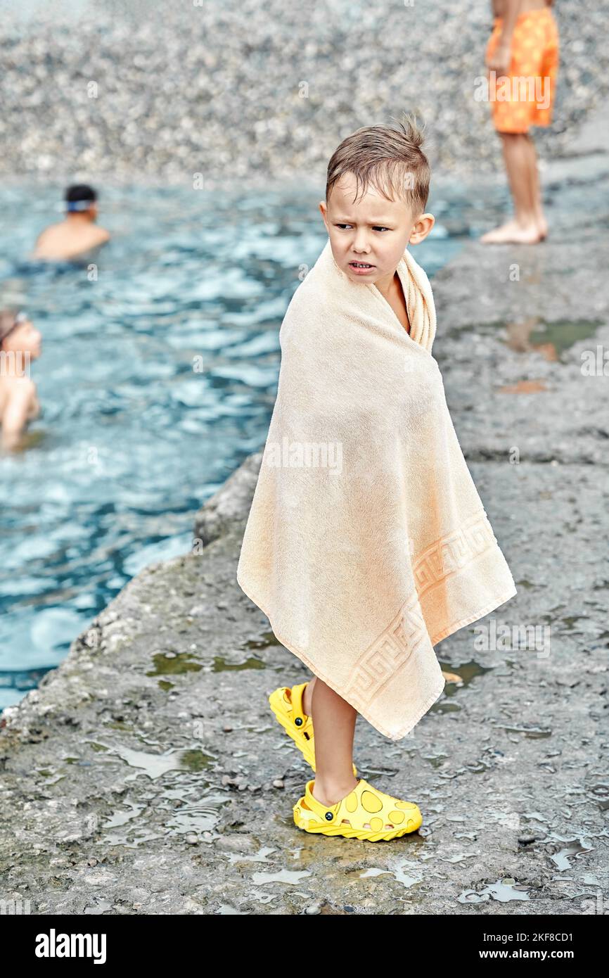 Der kleine Junge, der in ein beigefarbenes Frottee-Handtuch gehüllt ist, schaudert nach dem Schwimmen im Meer vor Kälte. Der Vorschuljunge versucht sich mit einem riesigen Handtuch aus der Nähe des Steinpiers aufzuwärmen Stockfoto
