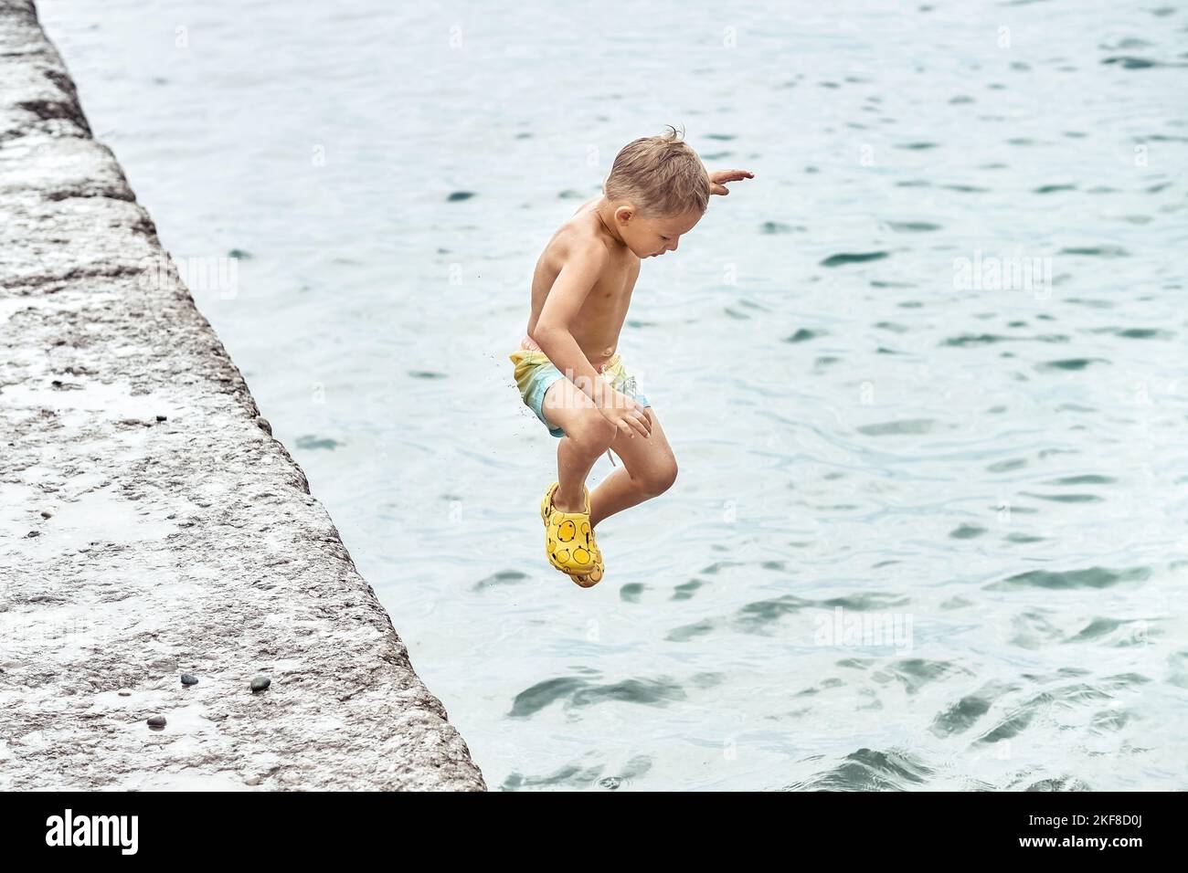 Der kleine Junge springt in lustiger Haltung vom Pier ins Meerwasser. Vorschulkinder verbringen gerne Urlaub mit der Familie am Meer Stockfoto