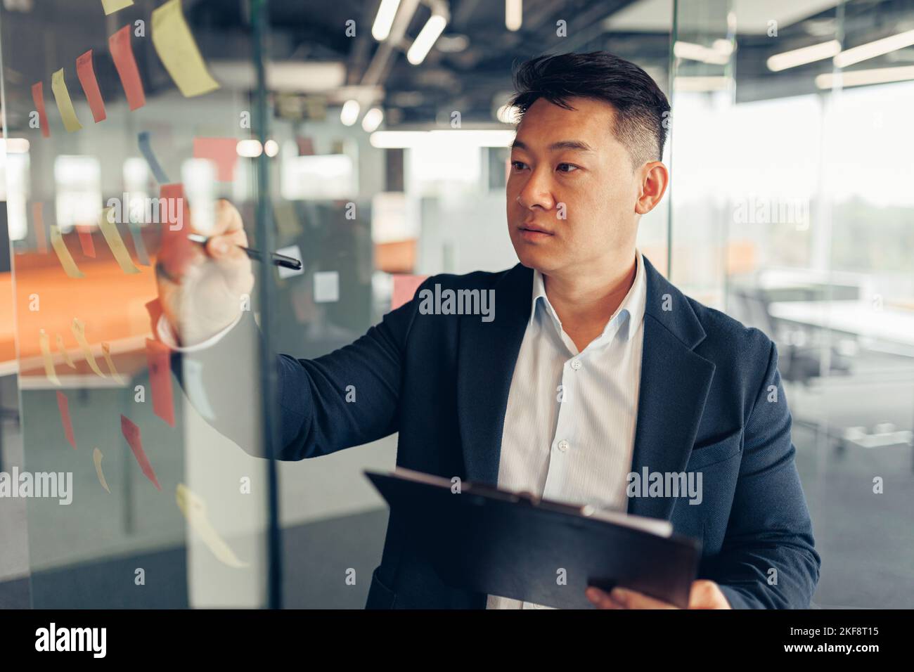 Fokussierter asiatischer Geschäftsmann, der auf Haftnotizen in der Glaswand des Büros schreibt Stockfoto