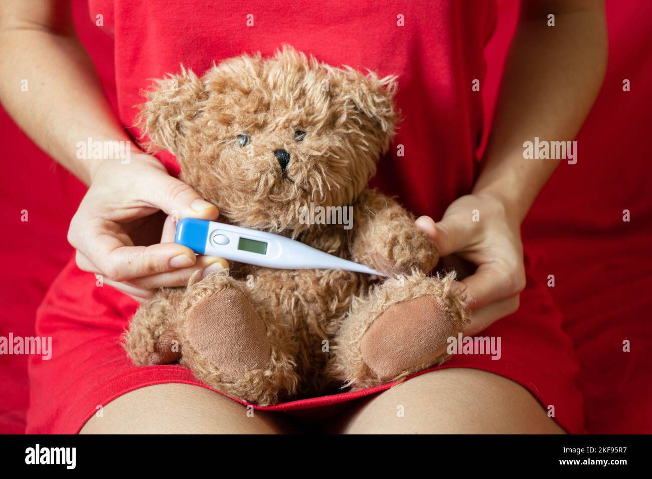 Mädchen hält einen braunen Teddybären mit einem Thermometer in der Hand zu Hause im Schlafzimmer auf dem Bett, Kindermorbidität, Fieber und Fieber, weltweit pand Stockfoto