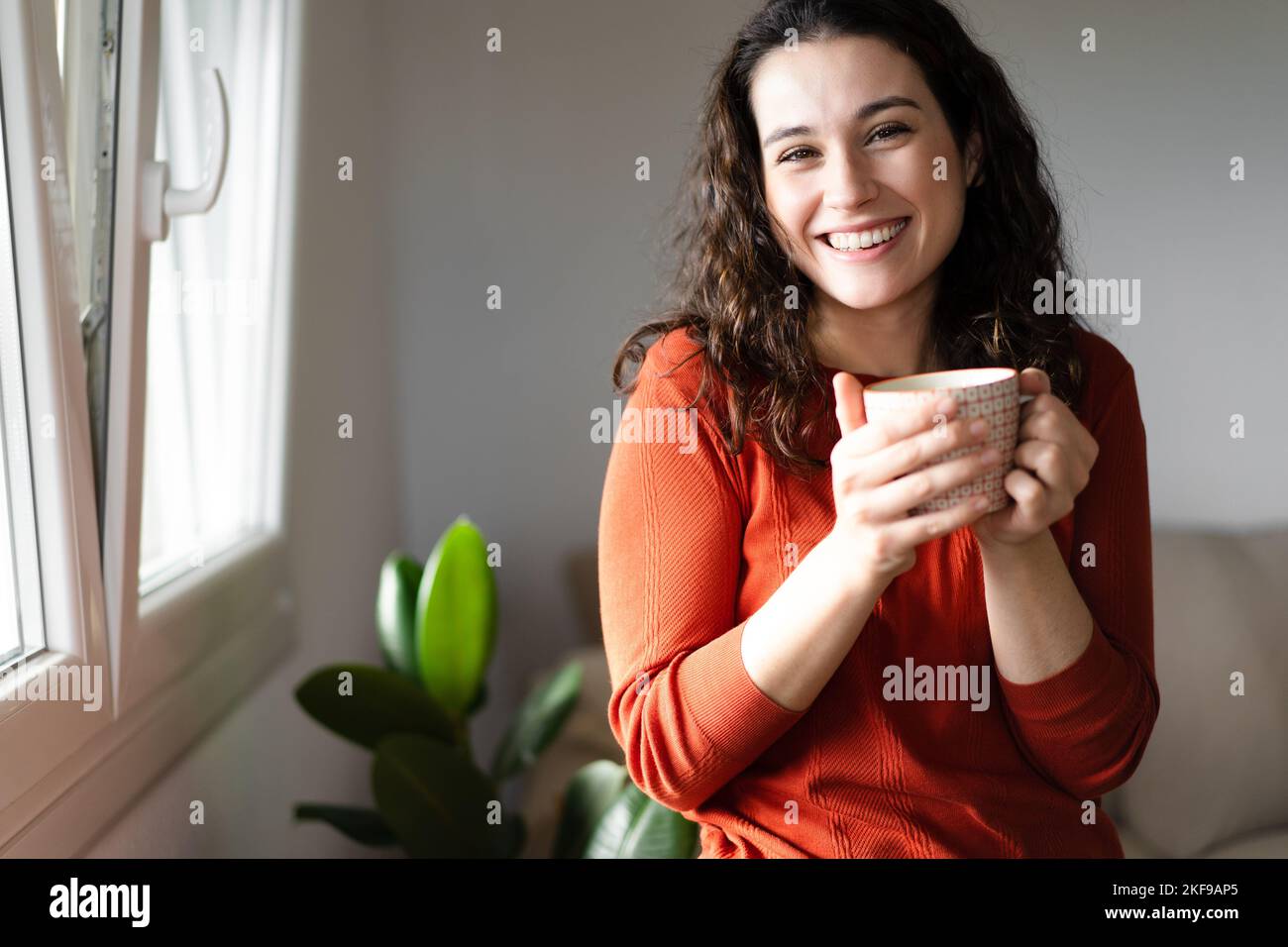 Junge, schöne Frau lacht, während sie zu Hause ein heißes Getränk am Fenster trinkt Stockfoto