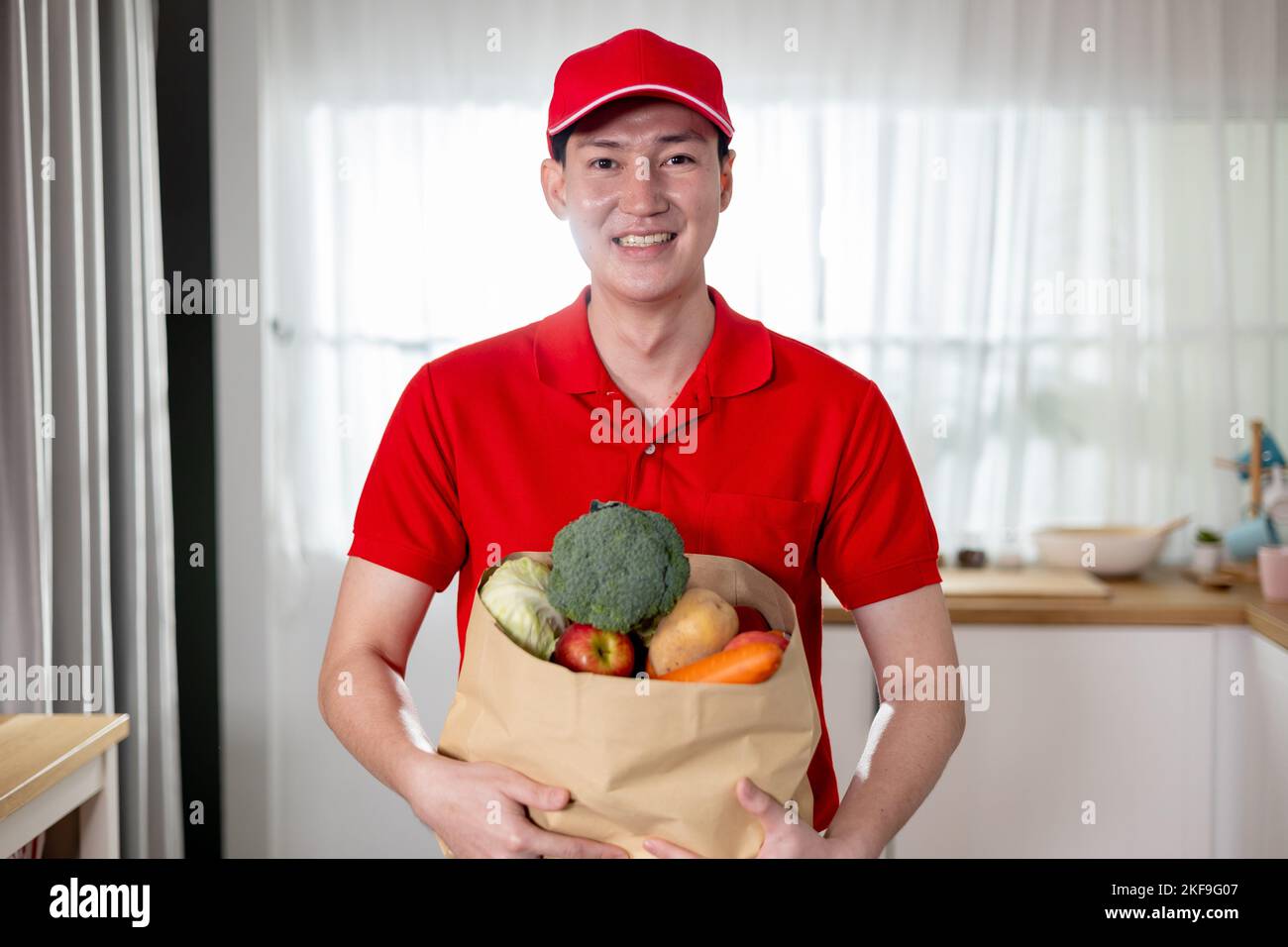 Lächelnder asiatischer Lieferer in roter Uniform, der Lebensmittel in einer Papiertüte trägt und Lebensmittel, Obst und Gemüse zu Hause liefert Stockfoto