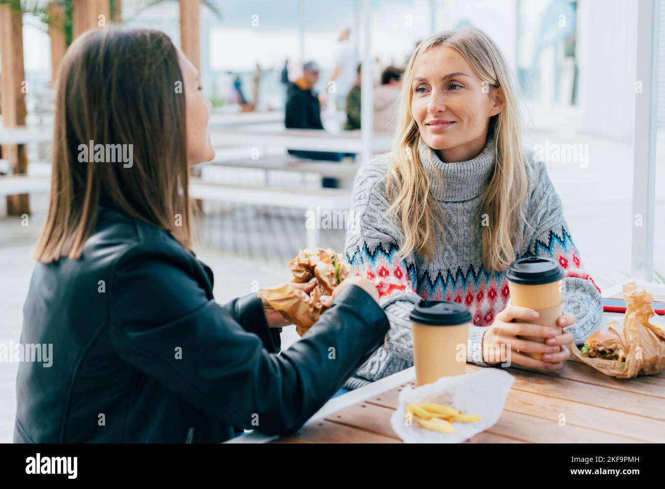 Zwei Freundinnen essen Burger, trinken Kaffee und plaudern auf der Straße, sitzen am Tisch. Stockfoto