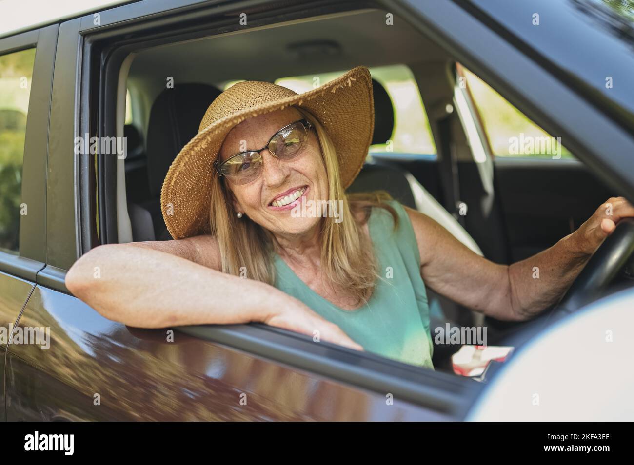 Fröhliche ältere Fahrerin im Strohhut, die in einem neuen Auto sitzt und lächelt und die Kamera anschaut, die die Reise genießt. Fahrkurse und Lebensversicherung. Aktivitäten im Ruhestand und Roadtrip-Konzept Stockfoto