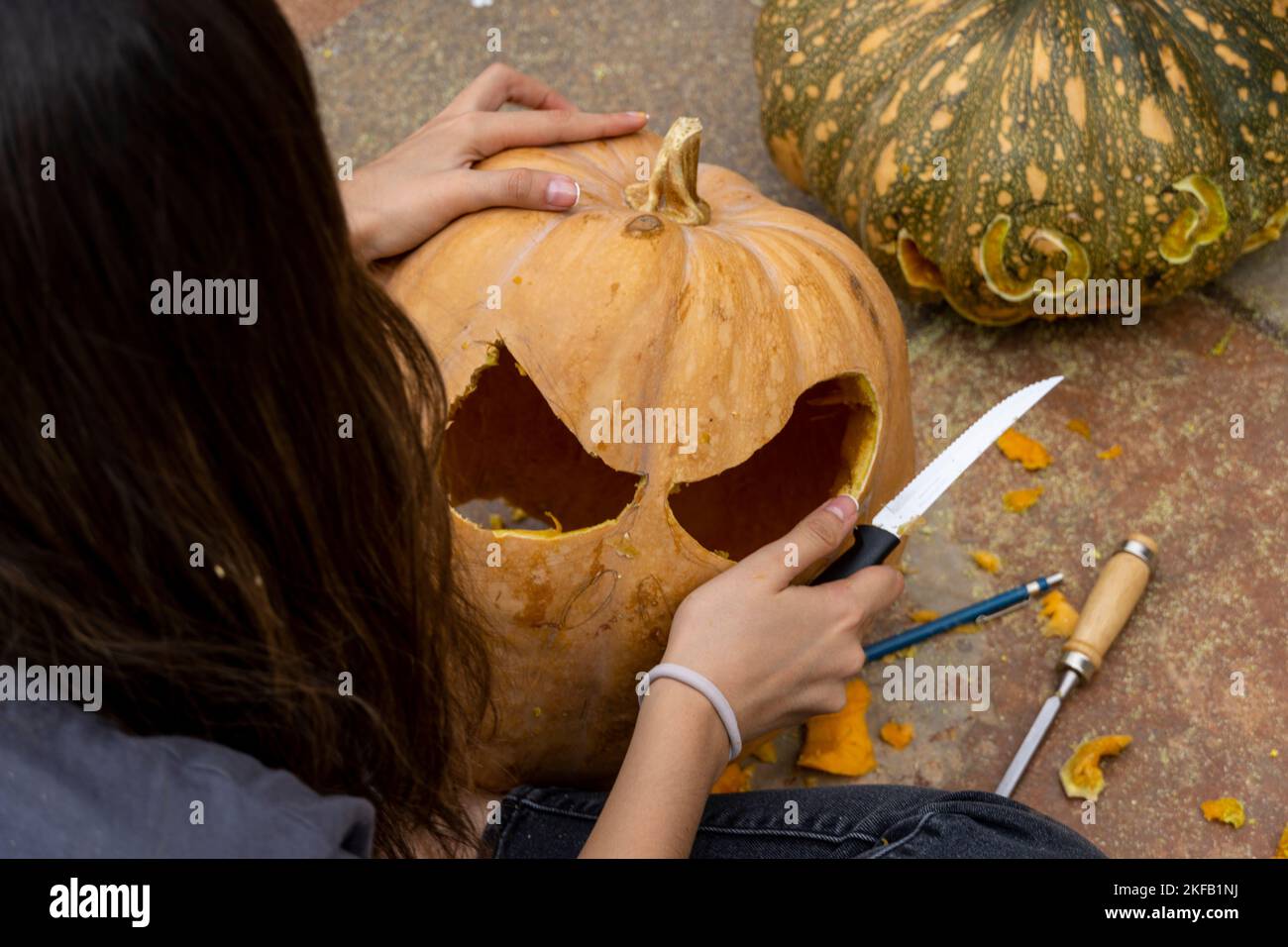 Frau schnitzt großen orangefarbenen Kürbis für Halloween, während sie zu Hause am Holztisch sitzt Stockfoto